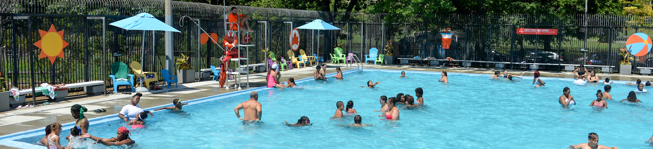 A busy outdoor city pool in a city park. There are brightly colored stencils of a sun and beach ball on the fence.