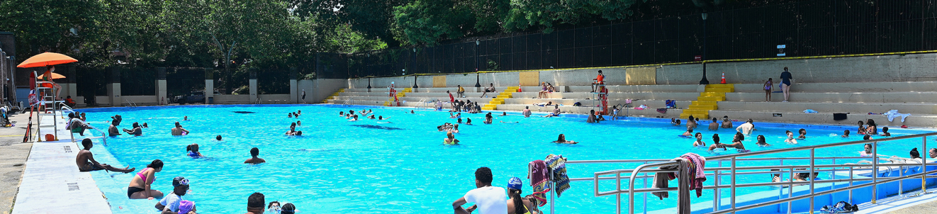 Children and adults relax in a large and tree-shaded outdoor swimming pool on a sunny day.