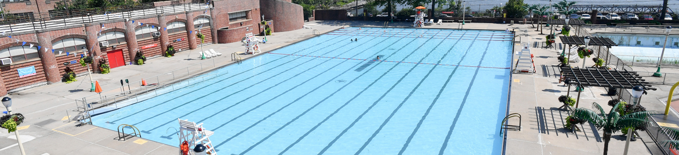 A large public pool and recreation complex located on the waterfront. The New York City skyline is visible in the distance across the bay.