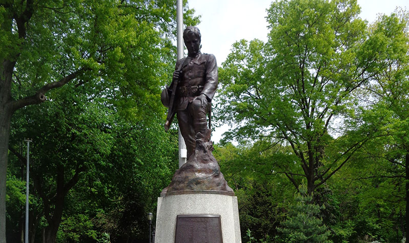 A bronze statue of a soldier with head bowed in contemplation of a small cross protruding from a mound of dirt.