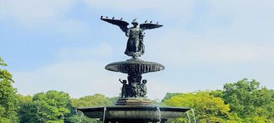 A figure of an angel stands atop the very tall Bethesda Fountain surrounded by trees in Central Park. Water runs down from the feet of the angel which was sculpted by lesbian sculptor Emma Stebbins.