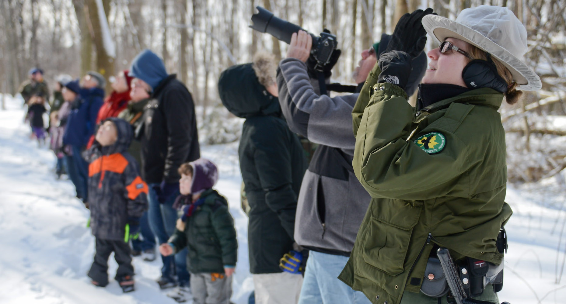 a park ranger and guests use binoculars to look to the trees for owls in a park covered in snow