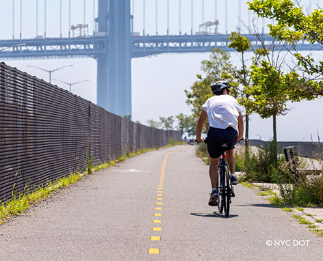 A cyclist rides on an asphalt path next to a fence and trees with a large bridge in the background.