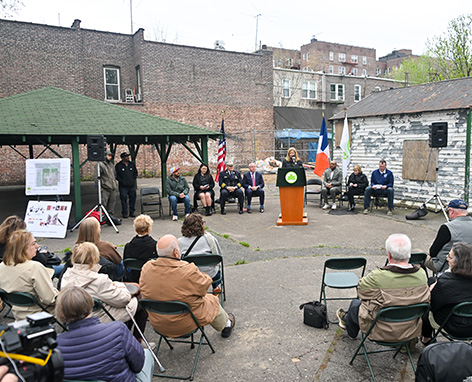 Parks Commissioner Sue Donahue stands behind a podium addressing an audience of seated community members gathered in a park.