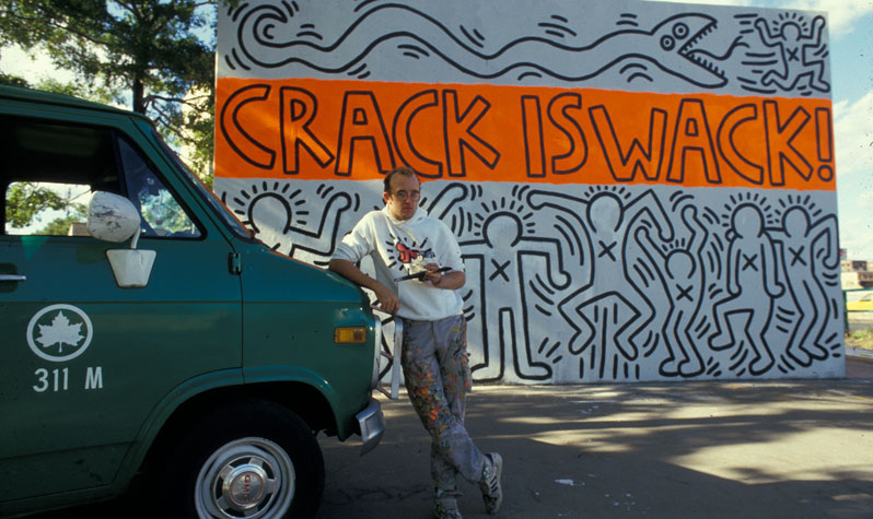 Keith Haring leans on a green parks van in front of his famous Crack is Wack mural, and orange and white artwork featuring chalk-like outlines of people with Xs on their chests.