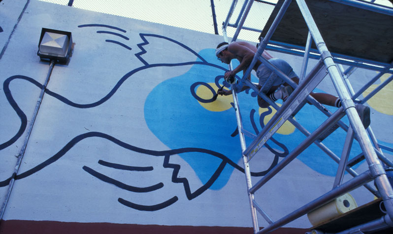 Keith Haring uses a scaffold to paint an outline of a fish with blotches of yellow and blue on the wall of the outdoor pool in what is now Tony Dapolito Park.